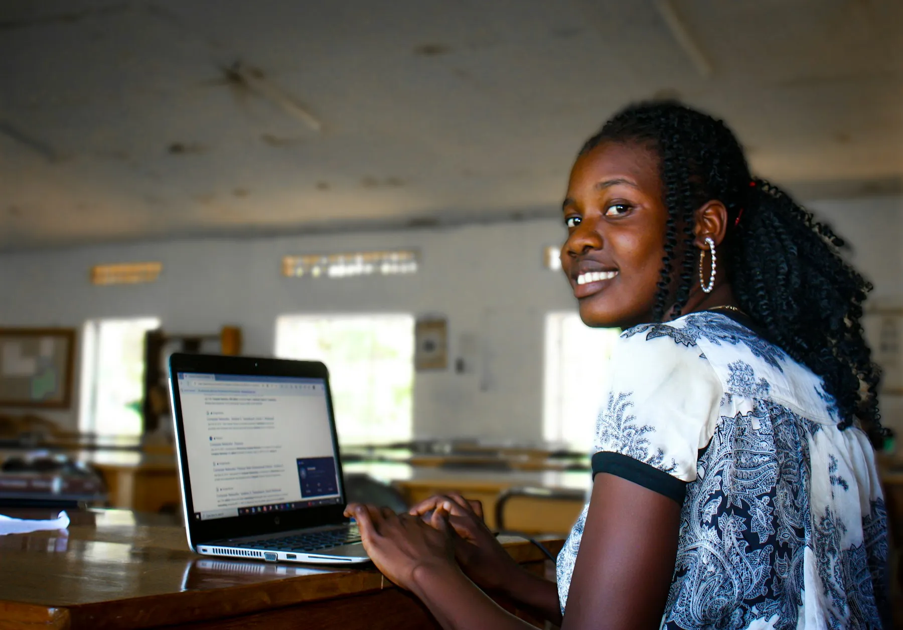 A young woman smiles over her shoulder while working on a laptop in a reading hall