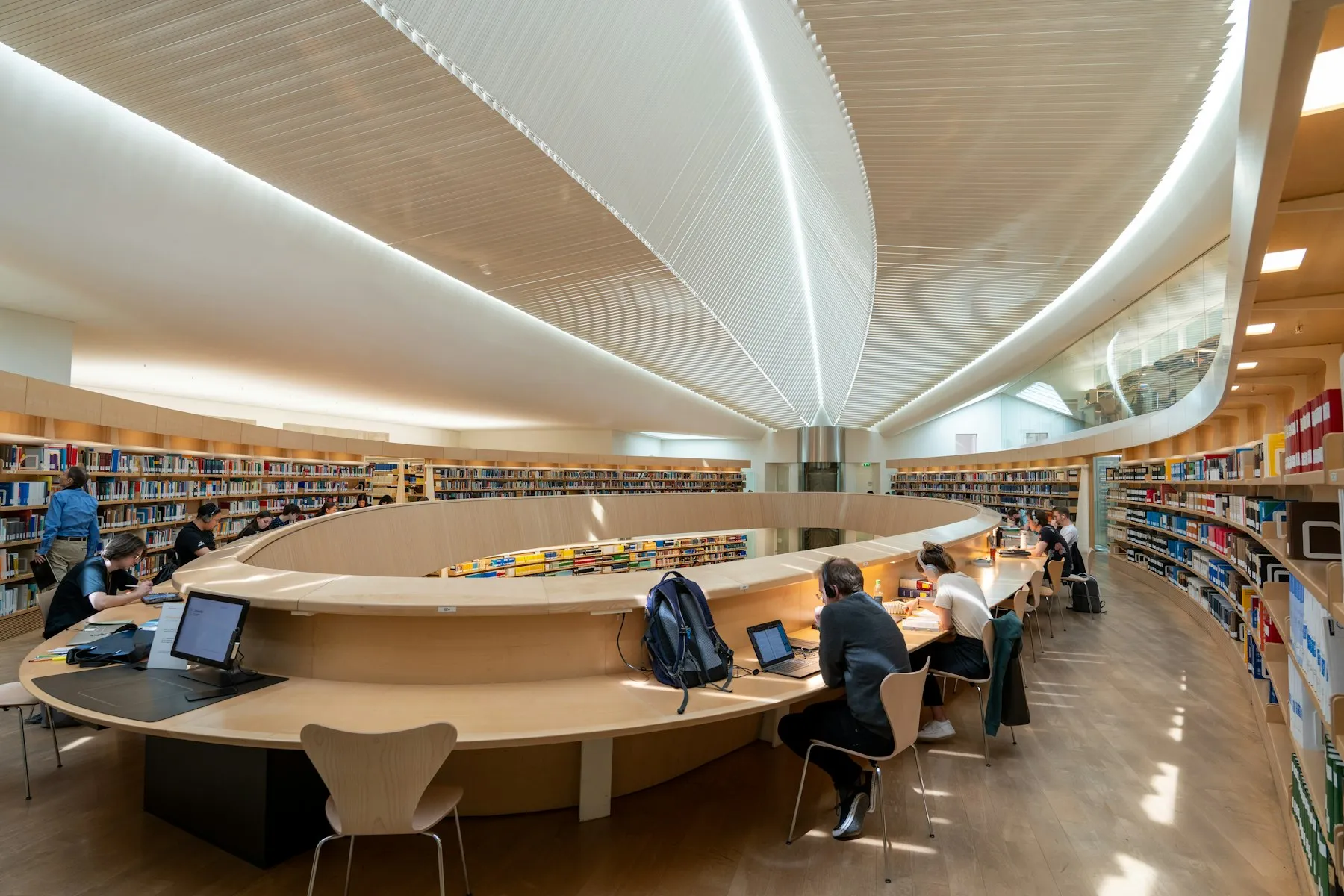 Readers at tables in a busy library, surrounded by shelves of books
